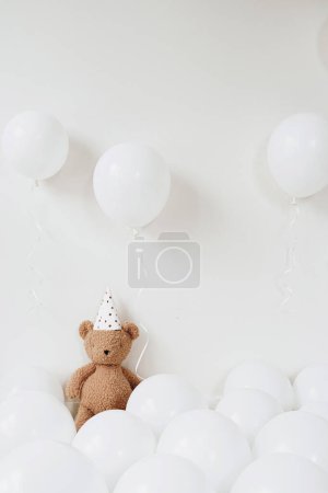 Téléchargez les photos : Ours en peluche en chapeau de fête assis près de ballons sur fond blanc - en image libre de droit