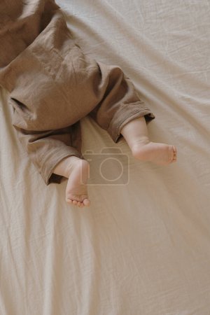 Photo for Cute newborn baby in brown bodysuit lying on the bed with beige bed linens. Cute minimal baby fashion concept. Top view - Royalty Free Image