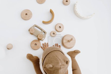Téléchargez les photos : Vue du dessus du bébé mignon d'un an en chapeau brun joue avec des jouets en bois élégants. Minimal boutique en ligne de mode bébé, concept de boutique en ligne - en image libre de droit