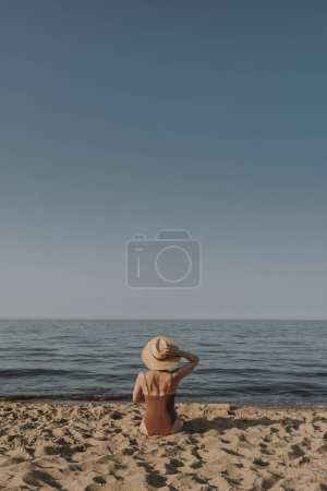 Foto de Hermosa mujer en sombrero de paja y traje de baño sentado en la arena en la playa y mirando en el mar y el cielo. Concepto estético de moda de verano - Imagen libre de derechos