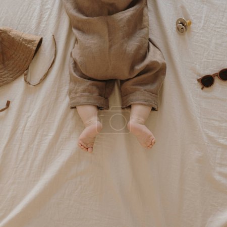 Foto de Bebé recién nacido en un elegante body básico con accesorios en ropa de cama de color beige neutro. Moda Ropa escandinava para recién nacidos. Sombrero, chupete, gafas de sol. Piso tendido, vista superior - Imagen libre de derechos
