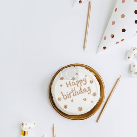 Téléchargez les photos : Gâteau d'anniversaire élégant avec signe "Joyeux anniversaire", bougies, cônes de fête sur fond blanc. Concept de célébration esthétique des fêtes. Couché à plat, vue de dessus. Couleurs blanc et or - en image libre de droit