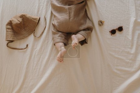 Photo for Cute newborn baby in brown bodysuit lying on the bed with beige bed linens. Cute minimal baby fashion concept. Top view - Royalty Free Image