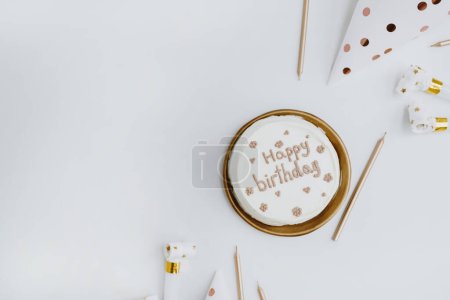 Téléchargez les photos : Gâteau d'anniversaire élégant avec signe "Joyeux anniversaire", bougies, cônes de fête sur fond blanc. Concept de célébration esthétique des fêtes. Couché à plat, vue de dessus. Couleurs blanc et or - en image libre de droit