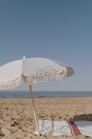 Téléchargez les photos : Minimal vacances d'été concept de vacances. Parapluie, serviette de plage sur sable devant le ciel bleu et la mer. Refroidissant, se prélasser sur la plage - en image libre de droit