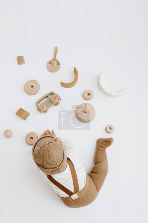 Téléchargez les photos : Petit bébé dans des vêtements élégants joue avec des jouets en bois. Mode Scandinave vêtements et jouets pour nouveau-nés. Chapeau, combinaison, combinaison. Couché plat, vue du dessus - en image libre de droit