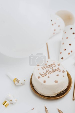 Foto de Elegante pastel de cumpleaños con signo "Feliz cumpleaños", velas, globos, conos festivos sobre fondo blanco. Concepto de celebración de eventos festivos estéticos. Colores blanco y dorado - Imagen libre de derechos