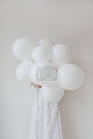 Photo for Woman in white dress holding bunch of white balloons over white wall. Aesthetic minimal festive holiday party event celebration. Balloons with blank copy space - Royalty Free Image