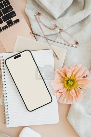 Photo for Blank smart phone screen with copy space. Comfortable home office workspace. Work at home. Laptop computer, notebook, poppy flower, earphones, glasses, cloth on table. Flat lay, top view - Royalty Free Image