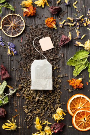 Foto de Composition of dry black and green tea, tea bag, dried citrus slices and dry hibiscus, calendula, lavender flowers on a dark wooden background top view close-up. Different types of dry tea on a dark background. - Imagen libre de derechos