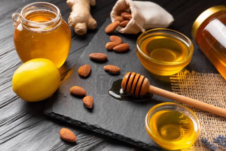 Honey in jars and a bowl, dipper, lemon, ginger, almond on a black stone plate on a wooden background. Conceptual composition of useful organic products. Natural products for the treatment of colds.