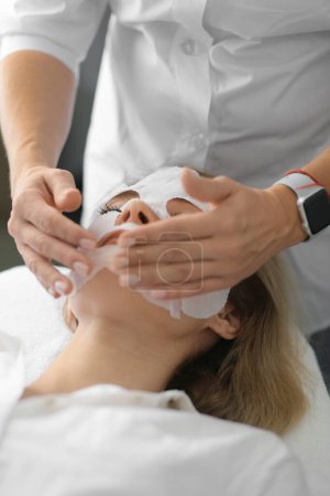 Téléchargez les photos : Beautician applies sheet mask on woman face for rehydrate face skin, anti aging cosmetic procedure in beauty spa salon. Cosmetologist applying moisturizing sheet mask on female face. Professional cosmetology. - en image libre de droit