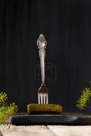 Photo for A pickled cucumber is pricked on a vintage fork on an aged dark board close-up. Homemade canned gherkin on a fork on a dark background. - Royalty Free Image