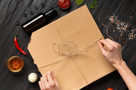 Photo for Pizza in a closed box, sauces and vegetables on a dark wooden background top view with space for text or advertising. Girl's hands open a box of pizza on the background of a dark table. Food delivery. - Royalty Free Image