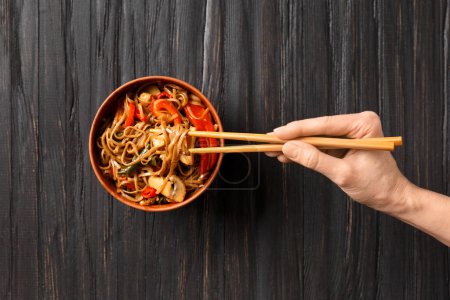 Photo for Plate with buckwheat noodles with vegetables, mushrooms, chicken meat and Chinese sticks on a dark background. Japanese soba in clay bowl on a wooden background. - Royalty Free Image