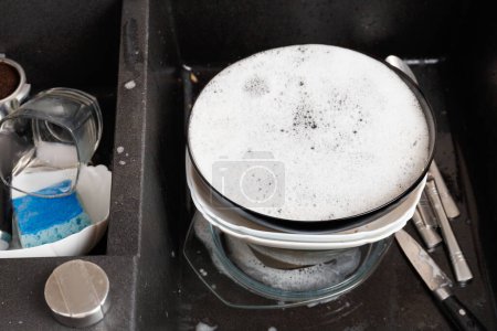 Foto de Concepto de lavar platos. Platos sucios con espuma de detergente en un fregadero de piedra negra en la vista superior de la cocina. Vasos sucios, tazas, platos, cubiertos en el fregadero de la cocina. - Imagen libre de derechos