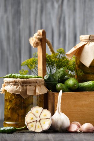 Téléchargez les photos : Fresh cucumbers in a wooden box, dill, garlic, chili peppers and pickled cucumber slices in a jar on a dark wooden background. Creative composition of fresh and pickled cucumbers spices and spices. - en image libre de droit