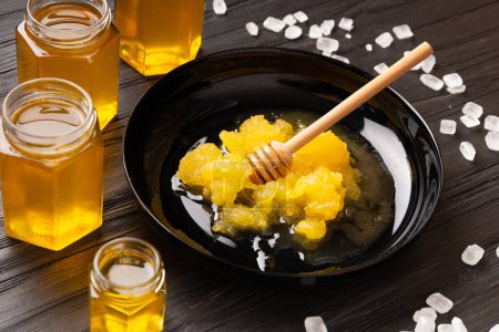 Foto de Honey in open jars, crystallized honey in a black plate and scattered sugar on a dark wooden background. Creative composition of different types of honey and large sugar crystals. - Imagen libre de derechos