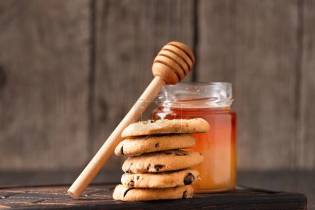 Foto de Honey in open jar, dipper and homemade cookies with chocolate chips on a wooden background. Homemade cakes and honey for breakfast. Composition of craft chocolate cookies and honey. - Imagen libre de derechos