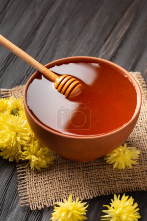 Foto de Miel en un bol de barro, cazo y flores sobre un fondo textil y de madera. Un palo para la miel se encuentra en un tazón de barro con miel de cerca. Composición de la miel en un plato y flores sobre un fondo oscuro - Imagen libre de derechos