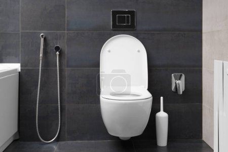 Foto de Moderno inodoro blanco montado en la pared, botón de color cromo y bidé ducha higiénica contra el fondo de una pared de baño negro. Parte del interior del baño en el apartamento. - Imagen libre de derechos