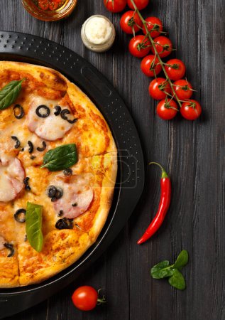 Photo for Pizza with meat , olives, cheese and herbs on a black dish, sauce, olive oil with spices and fresh vegetables on a dark wooden background top view. Traditional Italian food. - Royalty Free Image