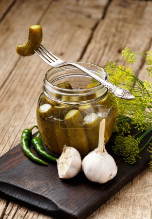 Photo for Pickled gherkins in a transparent glass jar, one canned cucumber on a vintage fork, fresh garlic, herbs and spices on a wooden background. - Royalty Free Image
