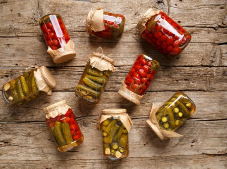 Téléchargez les photos : Canned cucumbers and tomato in glass jars on an old wooden background top view. Canned cherry tomatoes and carnishons in jars with kraft lids on a wooden background. - en image libre de droit