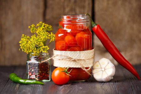 Photo for Pickled cherry tomatoes in an open glass jar, chili peppers, garlic, seasonings, spices and herbs for marinade close-up on a dark wooden background. Home preservation. - Royalty Free Image