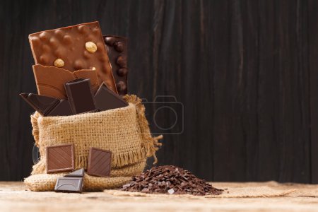Photo for Black and milk chocolate in a burlap bag, pieces of broken chocolate, chocolate chips on a wooden background with space for text. Handmade chocolate concept. - Royalty Free Image