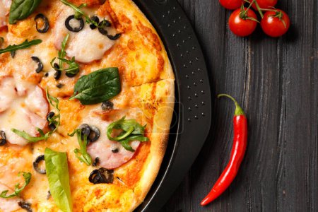 Photo for Pizza with jerky, olives, cheese and greens on a black round dish and fresh vegetables on a dark wooden background. Homemade pizza with meat and herbs. Traditional Italian food. - Royalty Free Image