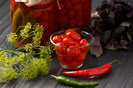 Photo for Pickled cherry tomatoes in a bowl, garlic, red and green chili peppers, dill, basil on a wooden background close-up, jars with canned tomatoes in the background. - Royalty Free Image
