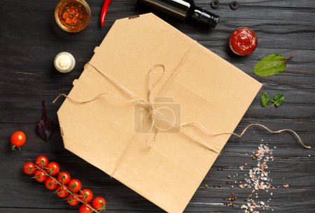 Foto de Pizza en una caja de cartón cerrada, salsas, aceite de oliva con especias, verduras frescas sobre un fondo de madera oscura vista superior. Entrega de alimentos. - Imagen libre de derechos