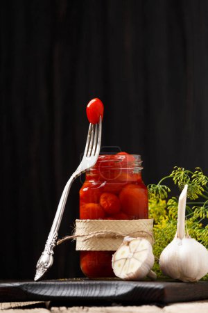 Téléchargez les photos : Tomates cerises marinées dans un bocal en verre ouvert, une tomate sur une fourchette vintage, ail entier et coupé en gros plan sur un fond en bois foncé. - en image libre de droit