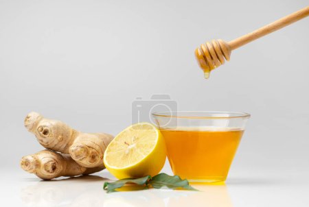 Foto de Miel en un tazón de vidrio, cazo vertiendo miel, jengibre y limón cortado con hojas verdes sobre un fondo blanco. Verter la miel en un frasco, palo de miel y cítricos de cerca en el aislamiento. - Imagen libre de derechos