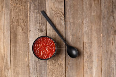 Photo for Red salmon caviar in an open black tin can and a black spoon on wooden background top view. Useful delicacy seafood, canned fish. - Royalty Free Image