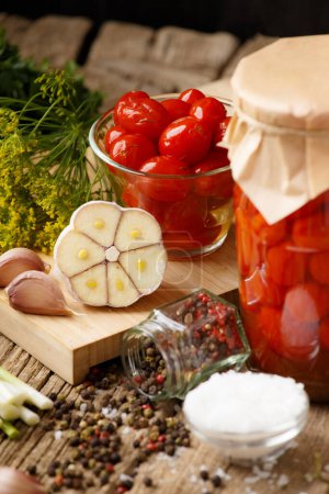 Téléchargez les photos : Tomates cerises marinées à l'ail, l'aneth et les épices. Tomates en conserve maison dans un bocal fermé avec un couvercle artisanal et dans un bol transparent sur un fond en bois. Légumes marinés. - en image libre de droit