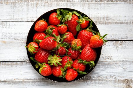Téléchargez les photos : Grandes baies juteuses mûres de fraises biologiques dans une assiette ronde noire sur fond bois clair, vue de dessus. - en image libre de droit