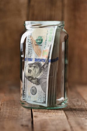 Foto de Billetes de dólar en un frasco de vidrio sobre un fondo de madera, una pila de billetes de 100 dólares en un frasco. El concepto de ahorrar dinero, acumular fondos. - Imagen libre de derechos