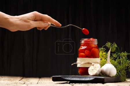 Photo for Pickled cherry tomatoes in an open transparent jar on a dark wooden background, one canned tomato on a vintage fork in a female hand. - Royalty Free Image