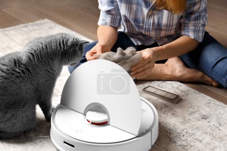 Téléchargez les photos : Le récipient du robot aspirateur est entre les mains des femmes, le collecteur de poussière du robot aspirateur est plein de laine, le chat est assis à proximité et regarde le récipient. - en image libre de droit