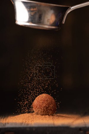 Photo for Cocoa powder is sprinkled on a chocolate truffle from a strainer under a beautiful light. A ball of chocolate truffle sprinkled with cocoa on a dark background close-up. - Royalty Free Image