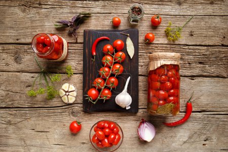 Téléchargez les photos : Tomates cerises en conserve dans des pots ouverts et fermés, ingrédients pour la conservation, une branche de tomates fraîches, poivrons, ail sur un fond en bois vieilli, vue sur le dessus. - en image libre de droit