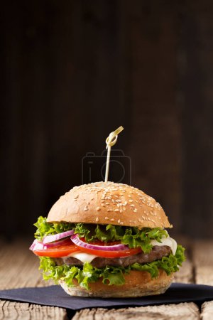 Téléchargez les photos : Hamburger appétissant avec escalope de bœuf juteuse, fromage, tomate, laitue, oignon sur une table en bois et fond sombre. Restauration rapide, hamburger maison. - en image libre de droit