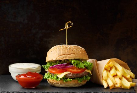 Téléchargez les photos : Hamburger de bœuf appétissant juteux, frites, deux sauces sur une assiette d'ardoise noire sur un fond sombre. Fast food américain, hamburger maison. - en image libre de droit