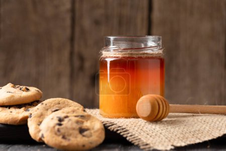 Téléchargez les photos : Honey in open jars and in a bowl, dipper and homemade cookies with chocolate chips on a wooden background. Homemade cakes and honey for breakfast. Composition of craft chocolate cookies and honey. - en image libre de droit