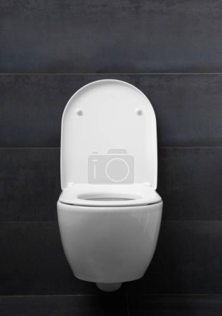 Photo for A modern wall-mounted white toilet with a lowered seat, a raised lid and a shiny chrome flush button against a black wall. Toilet bowl in the bathroom interior. - Royalty Free Image