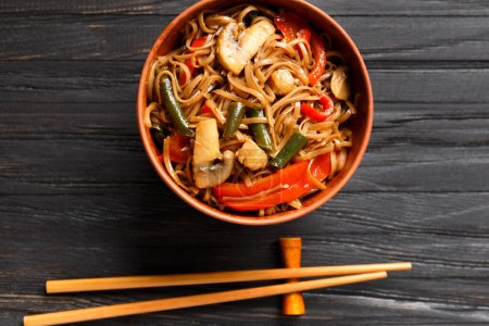 Photo for Plate with buckwheat noodles with vegetables, mushrooms, chicken meat, top view. Japanese soba in a clay bowl and Chinese chopsticks on a wooden background. - Royalty Free Image