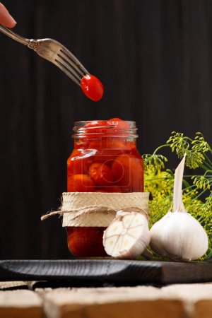 Foto de Tomates cherry en escabeche en un frasco abierto sobre una tabla de madera oscura, un tomate en un tenedor vintage sobre un frasco, fondo oscuro, primer plano. - Imagen libre de derechos