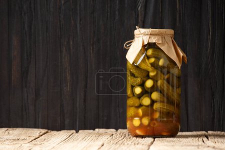 Photo for Pickled cucumbers in a closed glass jar with a craft lid on a wooden surface, dark background, copy space. - Royalty Free Image